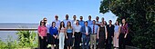Eine Gruppe von 14 Personen in halbformeller Kleidung versammelt sich vor dem Hintergrund eines ruhigen Gewässers und üppigen Grüns zu einem Foto.